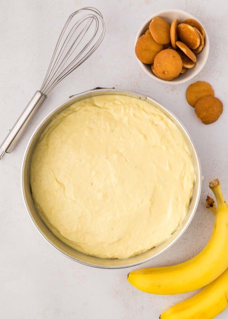 A mixing bowl of banana pudding with a whisk, a bunch of bananas, and a small bowl of vanilla wafers on a white surface.