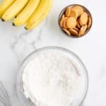A bunch of bananas, a bowl of vanilla wafers, a glass bowl of flour, and a whisk on a marble countertop.