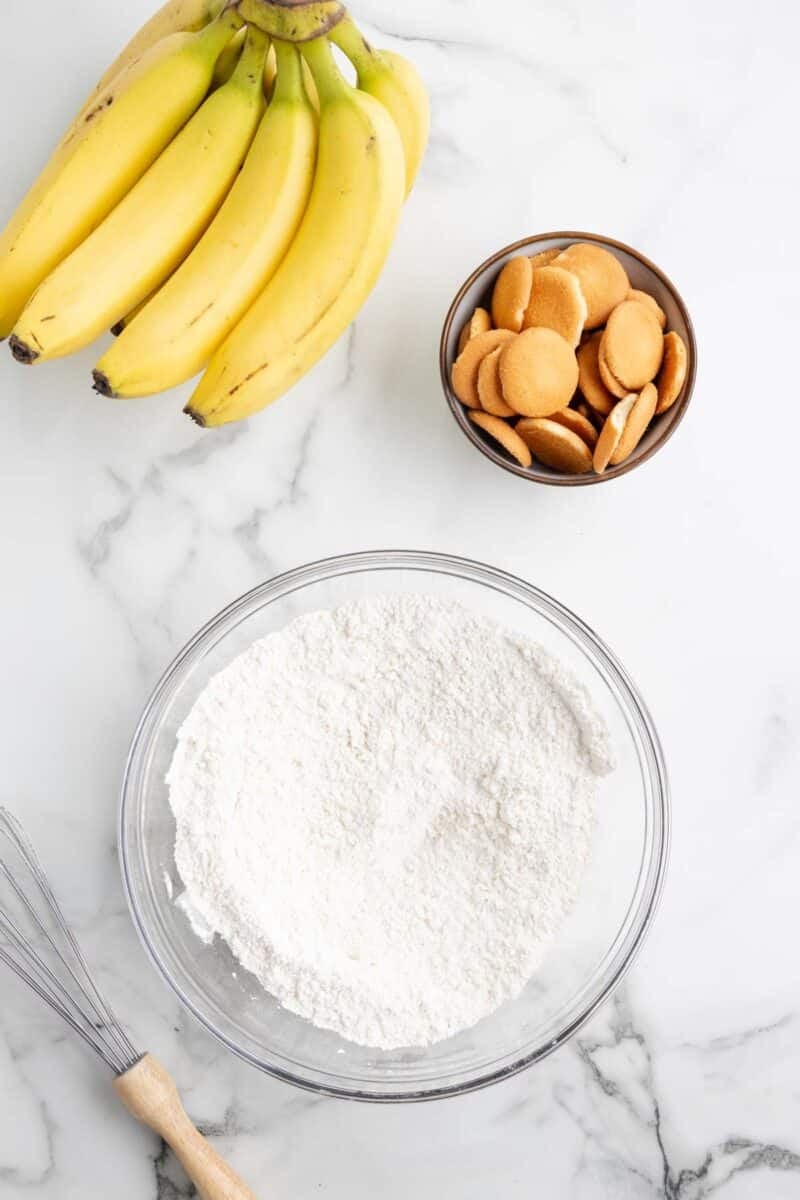 A bunch of bananas, a bowl of vanilla wafers, a glass bowl of flour, and a whisk on a marble countertop.