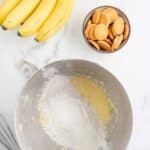 A bunch of bananas, a bowl of vanilla wafers, and a metal mixing bowl with flour and wet ingredients on a marble countertop, with a whisk nearby.