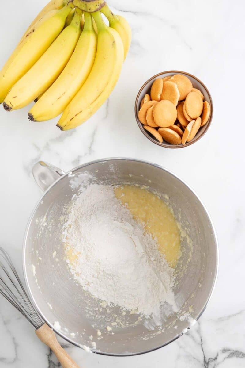 A bunch of bananas, a bowl of vanilla wafers, and a metal mixing bowl with flour and wet ingredients on a marble countertop, with a whisk nearby.