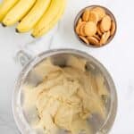 A bunch of bananas, a bowl of vanilla wafers, and a mixing bowl with banana pudding batter are arranged on a marble surface.