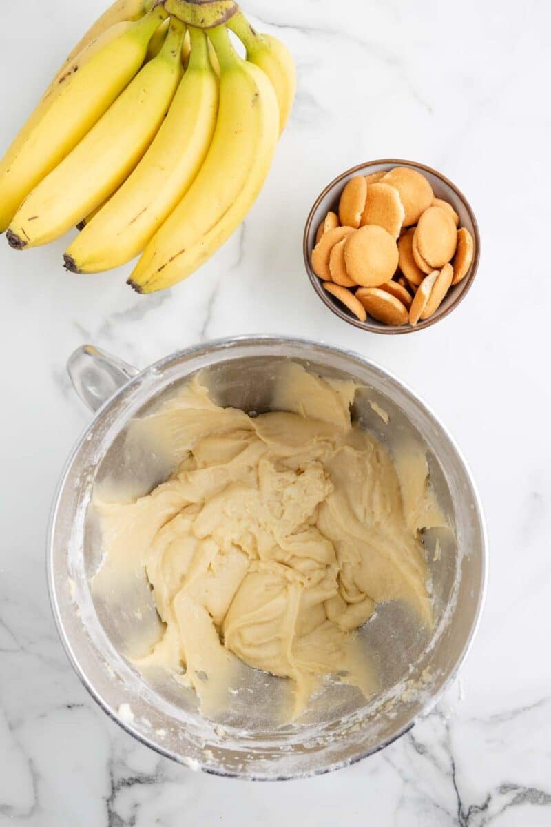 A bunch of bananas, a bowl of vanilla wafers, and a mixing bowl with banana pudding batter are arranged on a marble surface.