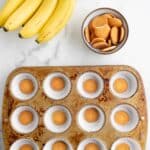 A muffin tin lined with paper cups, each containing a vanilla wafer, sits beside a bunch of bananas and a bowl of vanilla wafers on a marble surface.