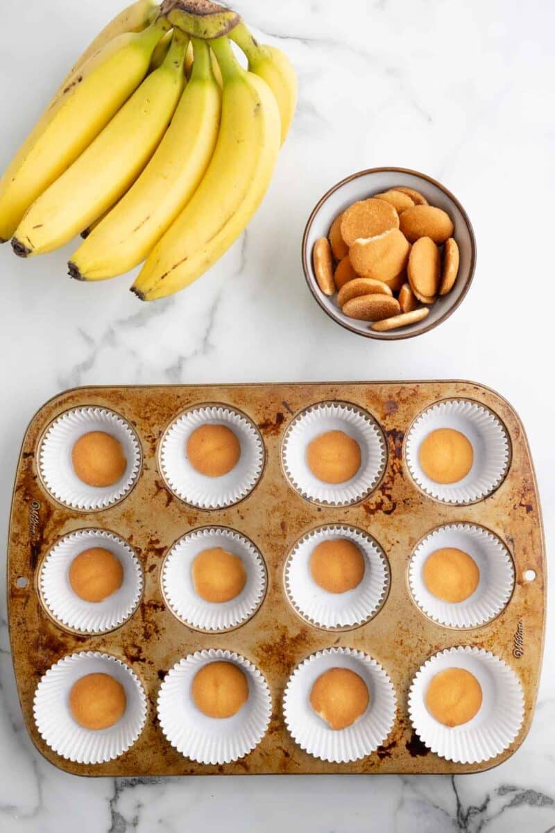 A muffin tin lined with paper cups, each containing a vanilla wafer, sits beside a bunch of bananas and a bowl of vanilla wafers on a marble surface.