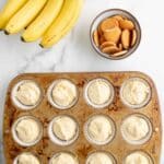 A bunch of bananas, a bowl of vanilla wafer cookies, and a muffin tin with twelve unbaked banana pudding cupcakes in paper liners are on a marble surface.