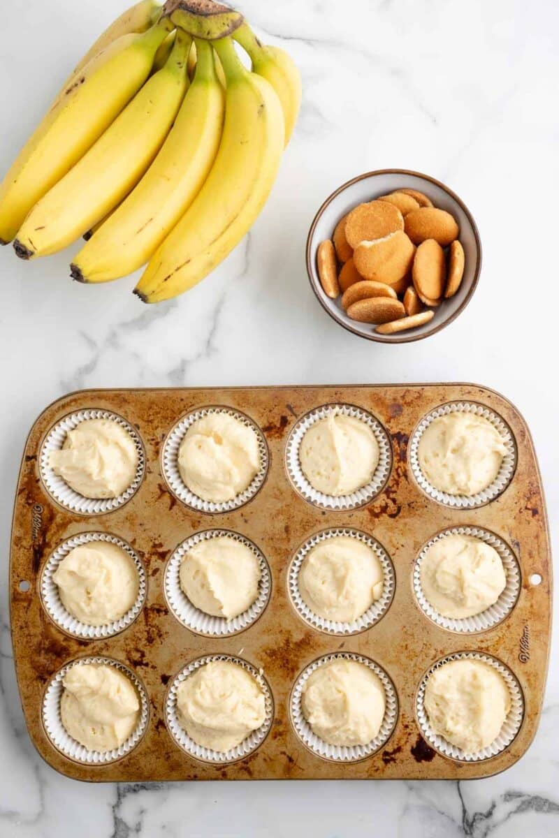 A bunch of bananas, a bowl of vanilla wafer cookies, and a muffin tin with twelve unbaked banana pudding cupcakes in paper liners are on a marble surface.
