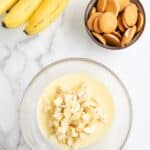 A bowl of chopped bananas in pudding mix sits on a marble surface next to a bunch of bananas, a bowl of vanilla wafers, and a whisk.