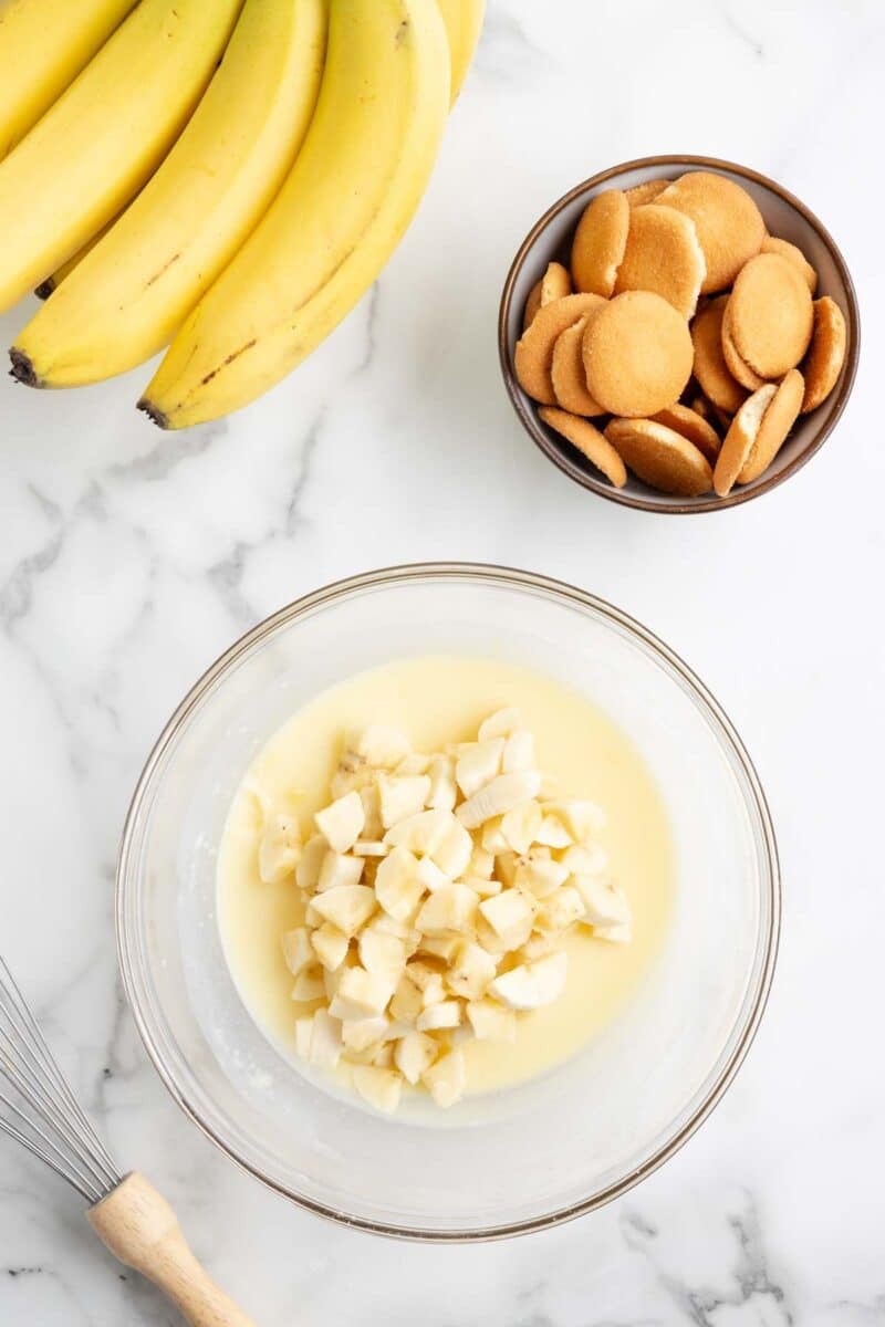 A bowl of chopped bananas in pudding mix sits on a marble surface next to a bunch of bananas, a bowl of vanilla wafers, and a whisk.