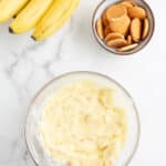A bunch of bananas, a bowl of vanilla wafer cookies, and a bowl of banana pudding on a marble countertop.