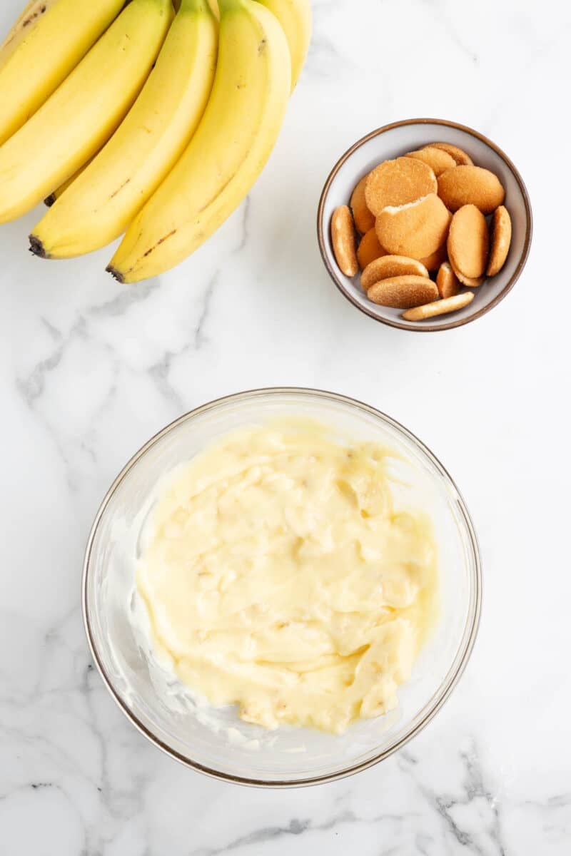 A bunch of bananas, a bowl of vanilla wafer cookies, and a bowl of banana pudding on a marble countertop.