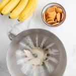 A bunch of bananas, a bowl of vanilla wafers, and a stand mixer bowl with cream cheese on a marble countertop.