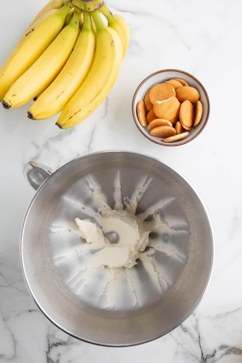 A bunch of bananas, a bowl of vanilla wafers, and a stand mixer bowl with cream cheese on a marble countertop.