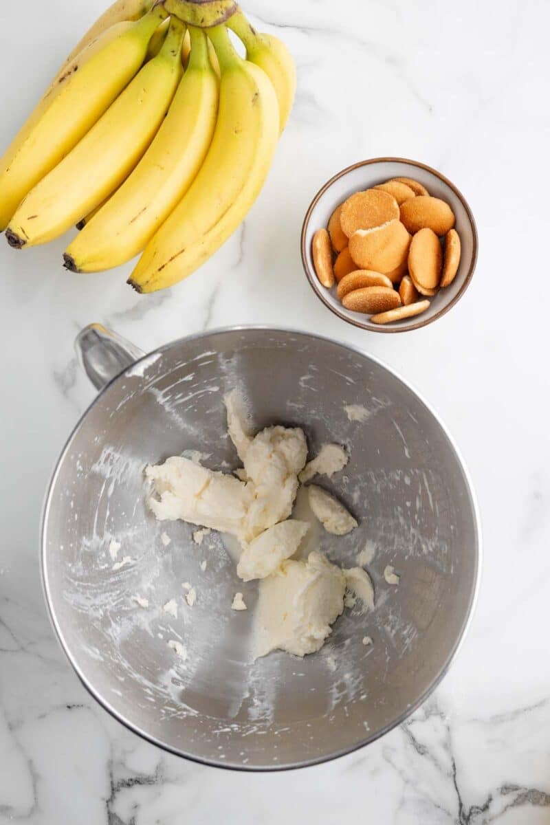 A bunch of bananas, a bowl of vanilla wafer cookies, and a metal mixing bowl with partially mixed butter and sugar on a marble countertop.