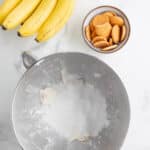 A bunch of bananas, a bowl of vanilla wafers, and a mixing bowl with powdered sugar on a marble countertop.
