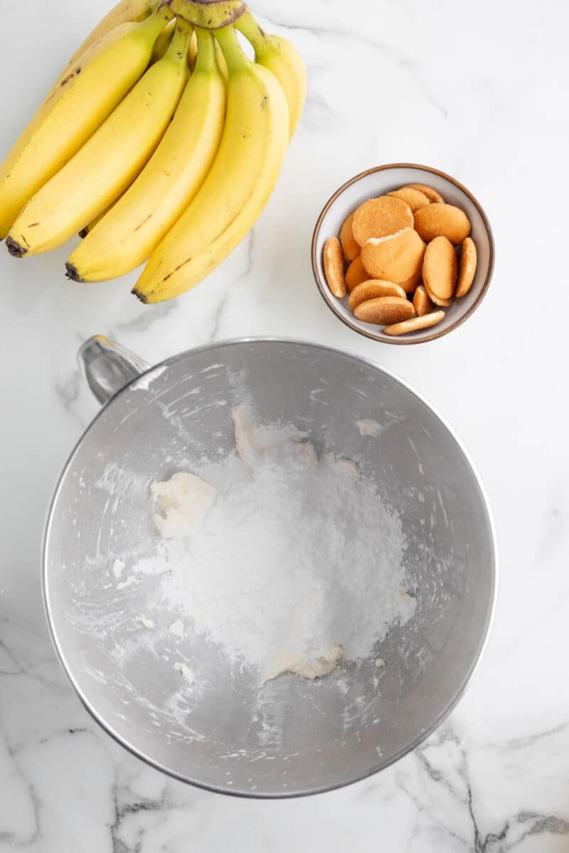 A bunch of bananas, a bowl of vanilla wafers, and a mixing bowl with powdered sugar on a marble countertop.