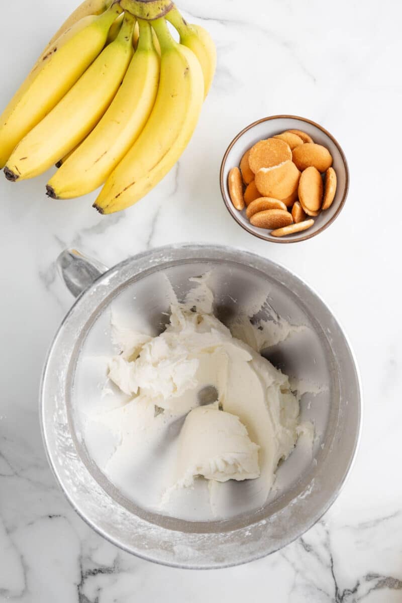 A bunch of bananas, a bowl of vanilla wafers, and a metal mixing bowl with whipped cream on a marble surface.