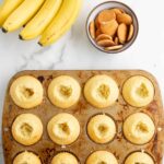 A muffin tin with cupcakes showing hollowed centers, a bunch of bananas, and a bowl of vanilla wafers on a marble surface.