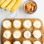A bunch of bananas, a bowl of vanilla wafers, and a muffin tin filled with cupcakes topped with swirled white frosting on a marble surface.