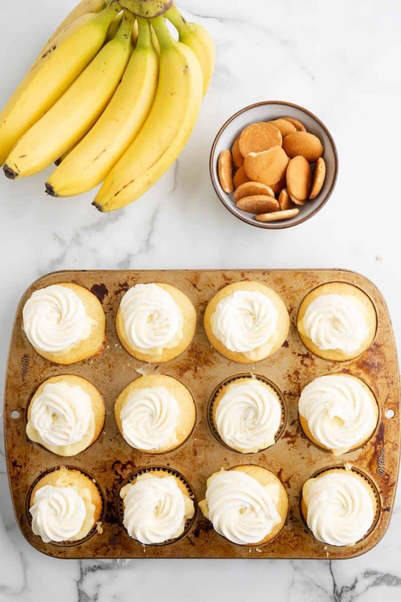 A bunch of bananas, a bowl of vanilla wafers, and a muffin tin filled with cupcakes topped with swirled white frosting on a marble surface.