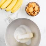A bunch of bananas, a bowl of vanilla wafers, and a metal mixing bowl with sugar and butter on a marble countertop.