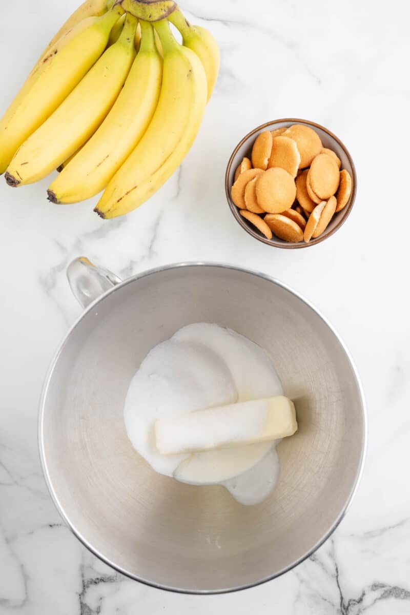 A bunch of bananas, a bowl of vanilla wafers, and a metal mixing bowl with sugar and butter on a marble countertop.