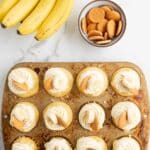 A muffin tin filled with cupcakes topped with whipped cream and vanilla wafers sits on a marble surface next to a bunch of bananas and a bowl of vanilla wafers.