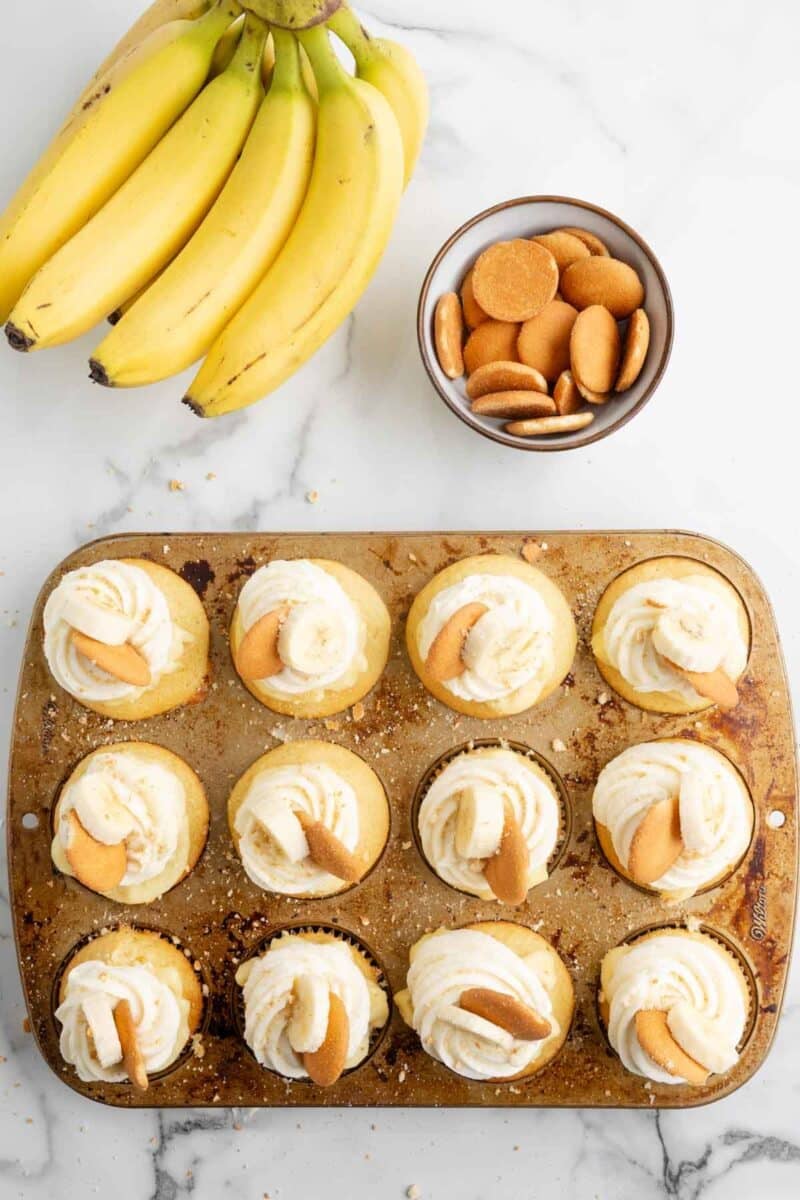 A muffin tin filled with cupcakes topped with whipped cream and vanilla wafers sits on a marble surface next to a bunch of bananas and a bowl of vanilla wafers.