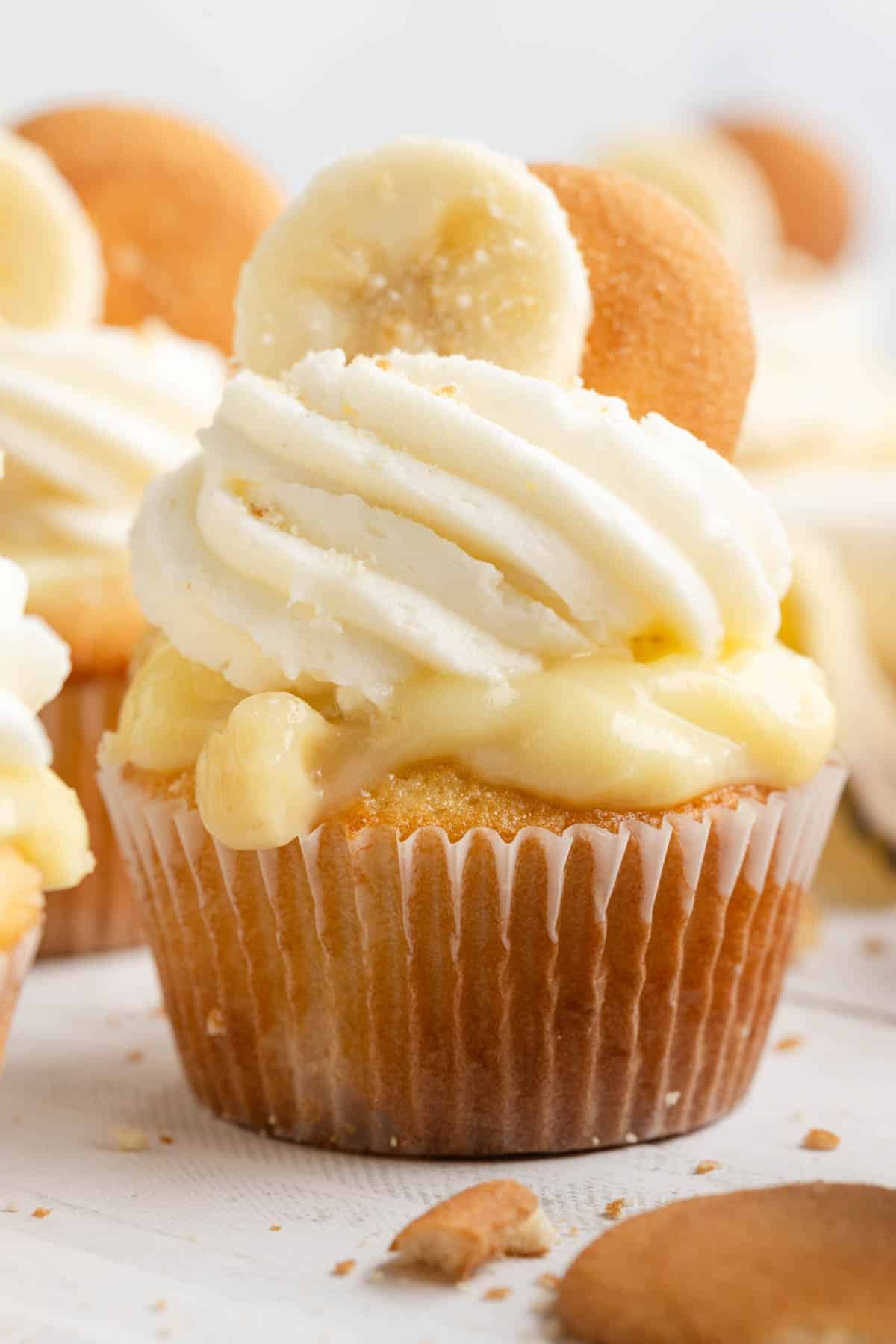 A banana pudding cupcake topped with swirled frosting, a banana slice, and a vanilla wafer, with custard filling visible underneath the frosting.