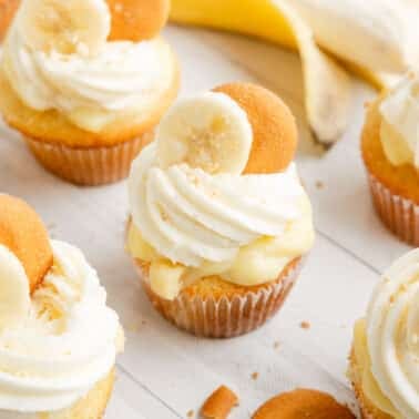 banana pudding cupcake topped with swirls of white frosting, banana slices, and vanilla wafers are arranged on a white surface with a peeled banana in the background.