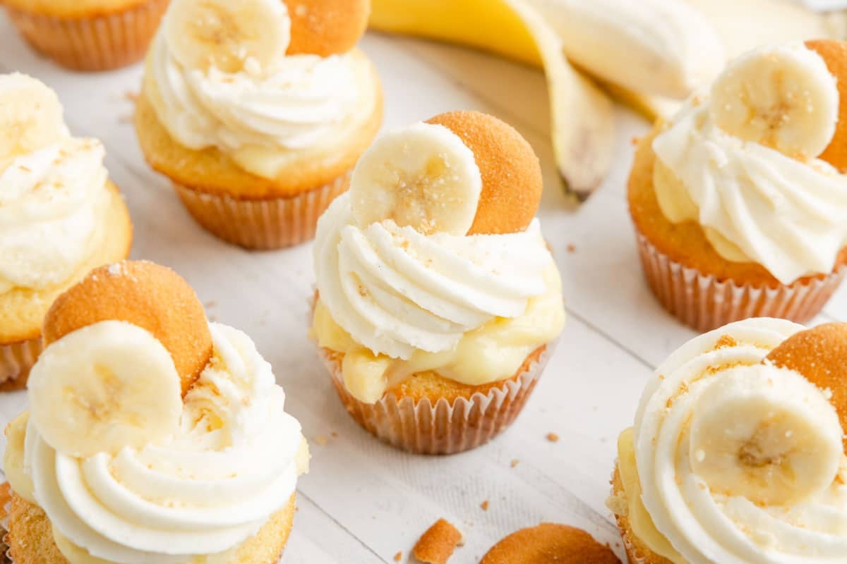 banana pudding cupcake topped with swirls of white frosting, banana slices, and vanilla wafers are arranged on a white surface with a peeled banana in the background.