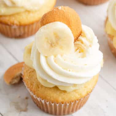 A banana pudding cupcake topped with whipped frosting, a banana slice, and a vanilla wafer, on a white wooden surface.