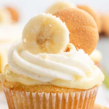 A banana pudding cupcake topped with swirled white frosting, a banana slice, and a vanilla wafer cookie.