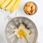 A mixing bowl with creamed butter and sugar and a cracked egg, next to a bunch of bananas and a bowl of vanilla wafer cookies on a marble surface.