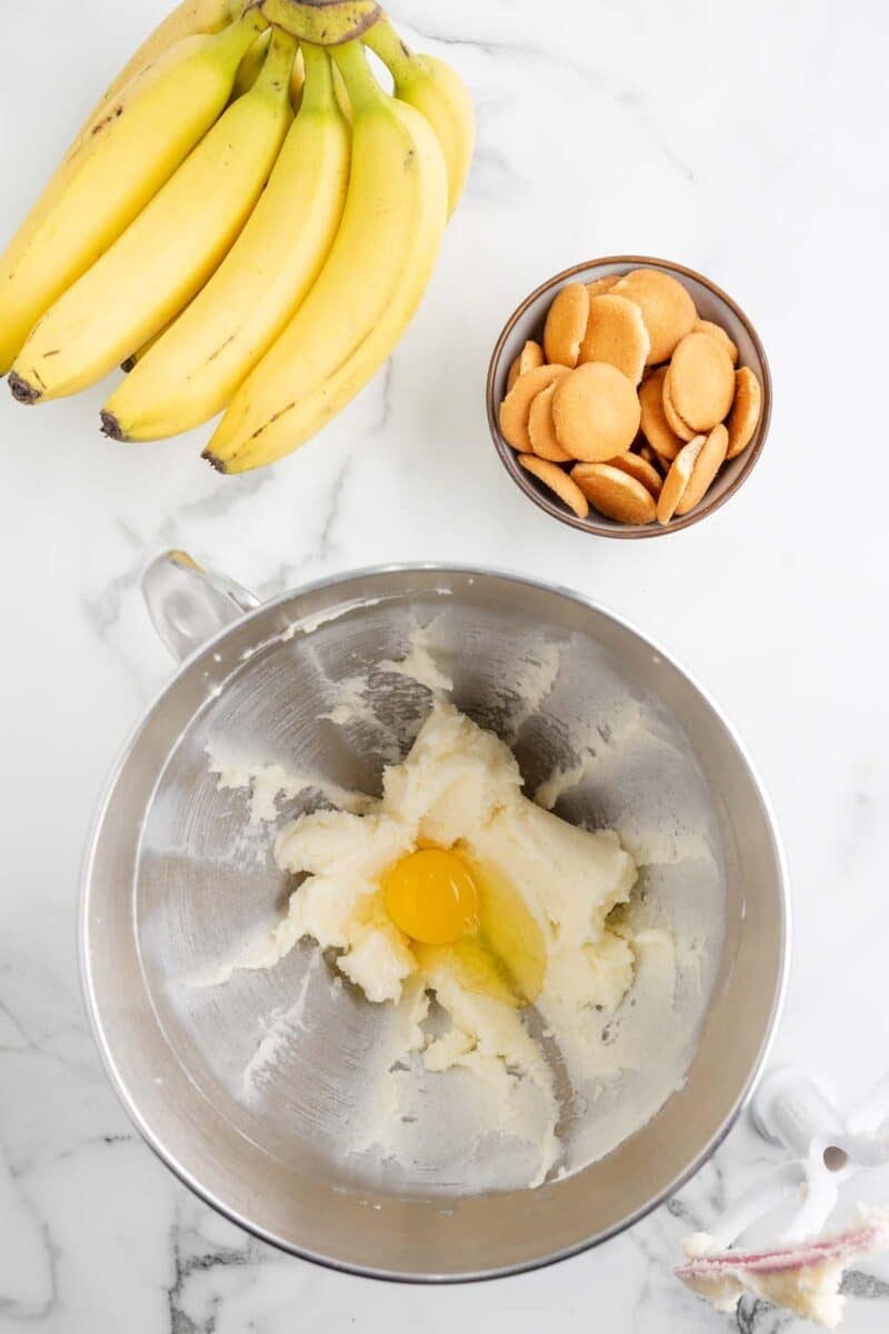 A mixing bowl with creamed butter and sugar and a cracked egg, next to a bunch of bananas and a bowl of vanilla wafer cookies on a marble surface.