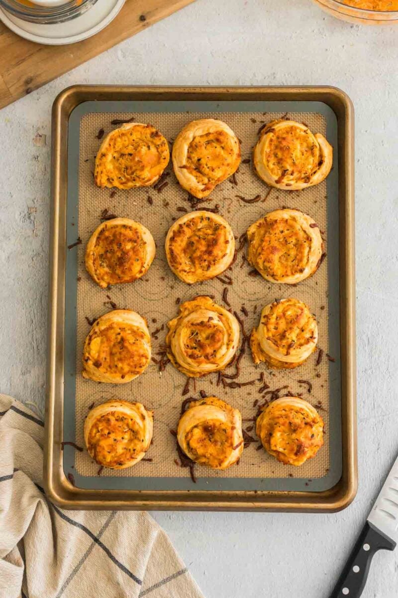 A baking sheet lined with parchment paper holds twelve evenly spaced, buffalo chicken pinwheels on a light-colored surface.