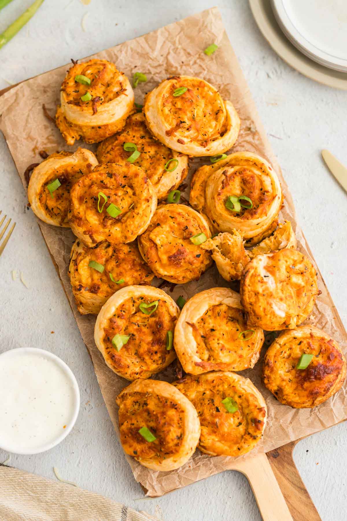 A wooden serving board with several buffalo chicken pinwheels, garnished with green onions, is placed on parchment paper next to a small bowl of white dipping sauce.