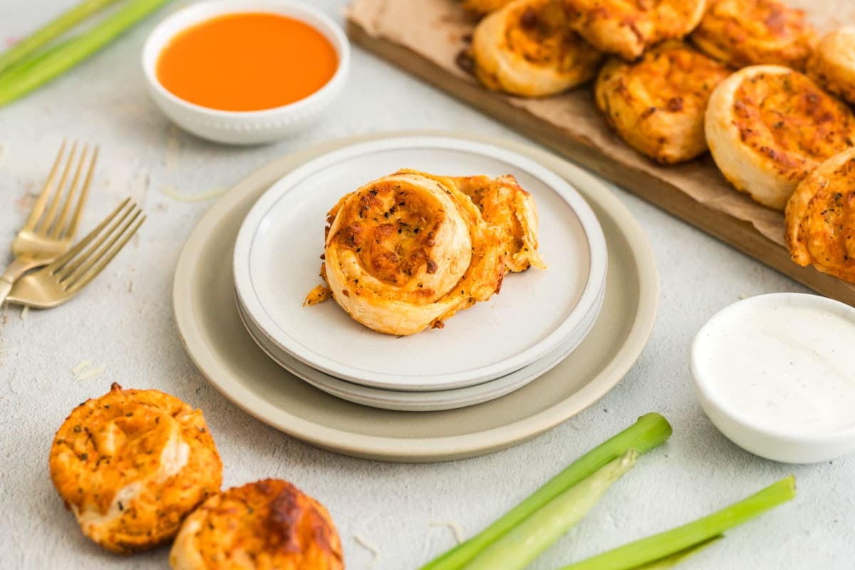 A plate with two puff pastry buffalo chicken pinwheels, surrounded by celery sticks, dipping sauces, and more pinwheels on parchment paper in the background.