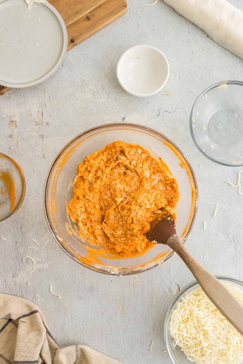 A glass bowl filled with a creamy orange mixture is being stirred with a spatula, surrounded by empty bowls and shredded cheese on a light-colored surface.