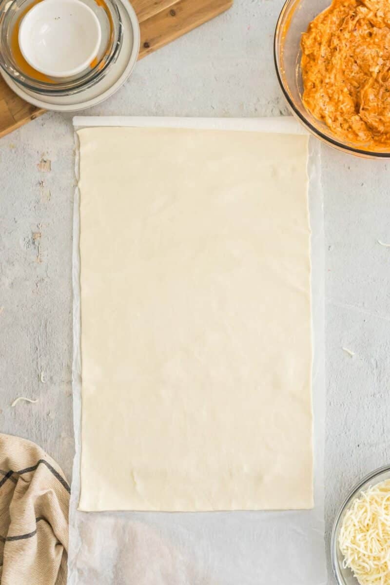 A sheet of puff pastry is laid out on parchment paper with bowls of creamy filling and grated cheese nearby on a light countertop.