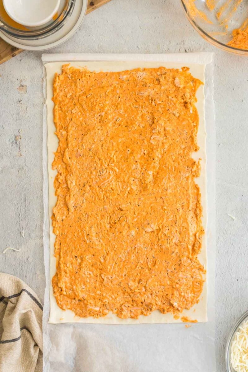 A rectangular sheet of dough topped with an orange, creamy spread is placed on parchment paper, surrounded by bowls and a cloth on a light surface.