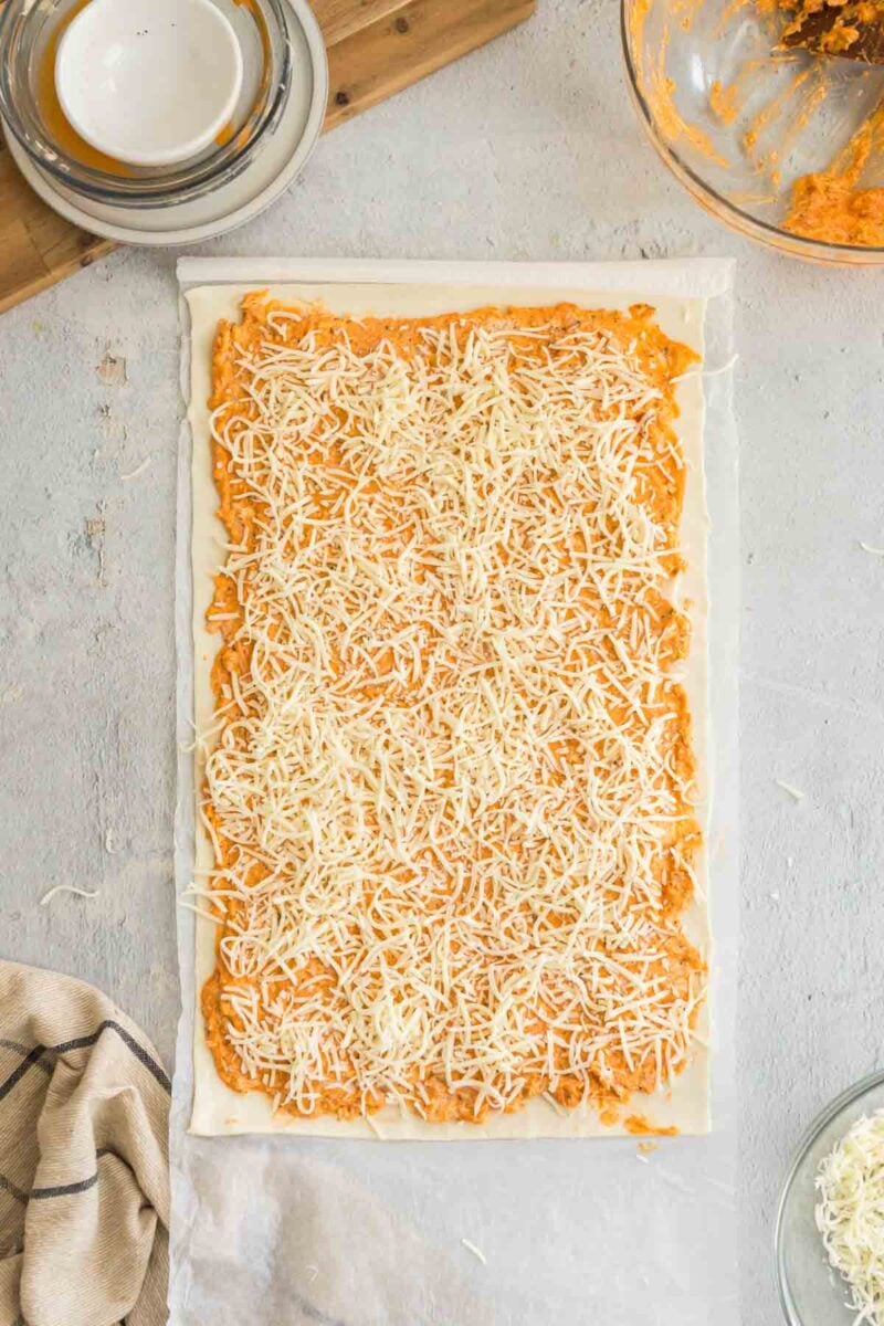 A rectangular sheet of dough topped with an orange spread and shredded cheese on a light surface, with bowls and a cloth nearby.