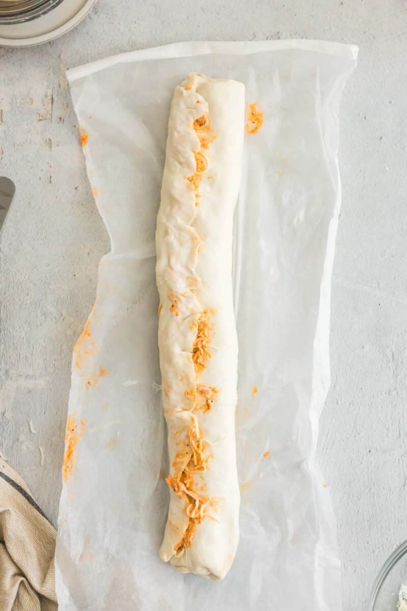 A rolled, unbaked pastry filled with shredded cheese sits on a sheet of parchment paper against a light-colored surface.
