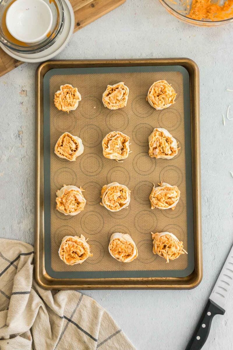A baking sheet with twelve unbaked pinwheels filled with shredded orange and white filling, placed on a light countertop with a towel, knife, and stack of plates nearby.