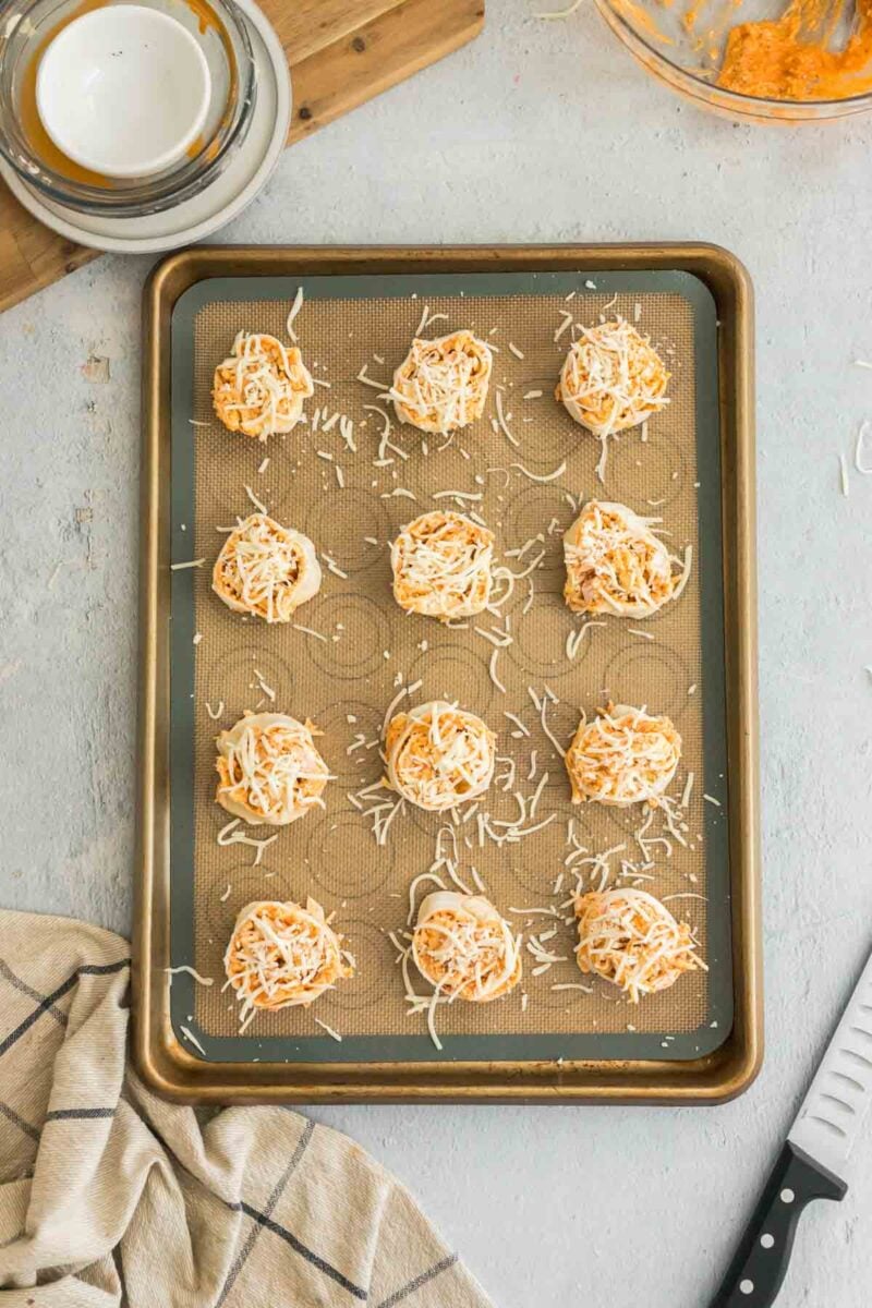Twelve uncooked cheesy balls are arranged on a baking sheet lined with a silicone mat, ready to be baked. A knife, towel, and bowl are nearby.