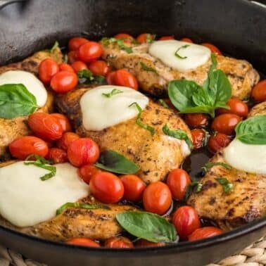 Caprese chicken skillet topped with melted cheese, fresh basil, and surrounded by cherry tomatoes.