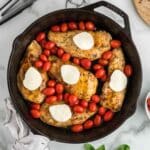 A cast iron skillet with cooked chicken breasts, cherry tomatoes, and slices of mozzarella cheese on a marble countertop. Tongs, a wooden board, a napkin, and fresh basil are nearby.