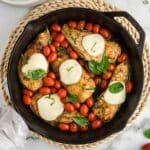 Caprese chicken skillet topped with melted cheese, cherry tomatoes, and fresh basil, placed on a woven placemat. Plates, forks, and grape tomatoes are nearby.