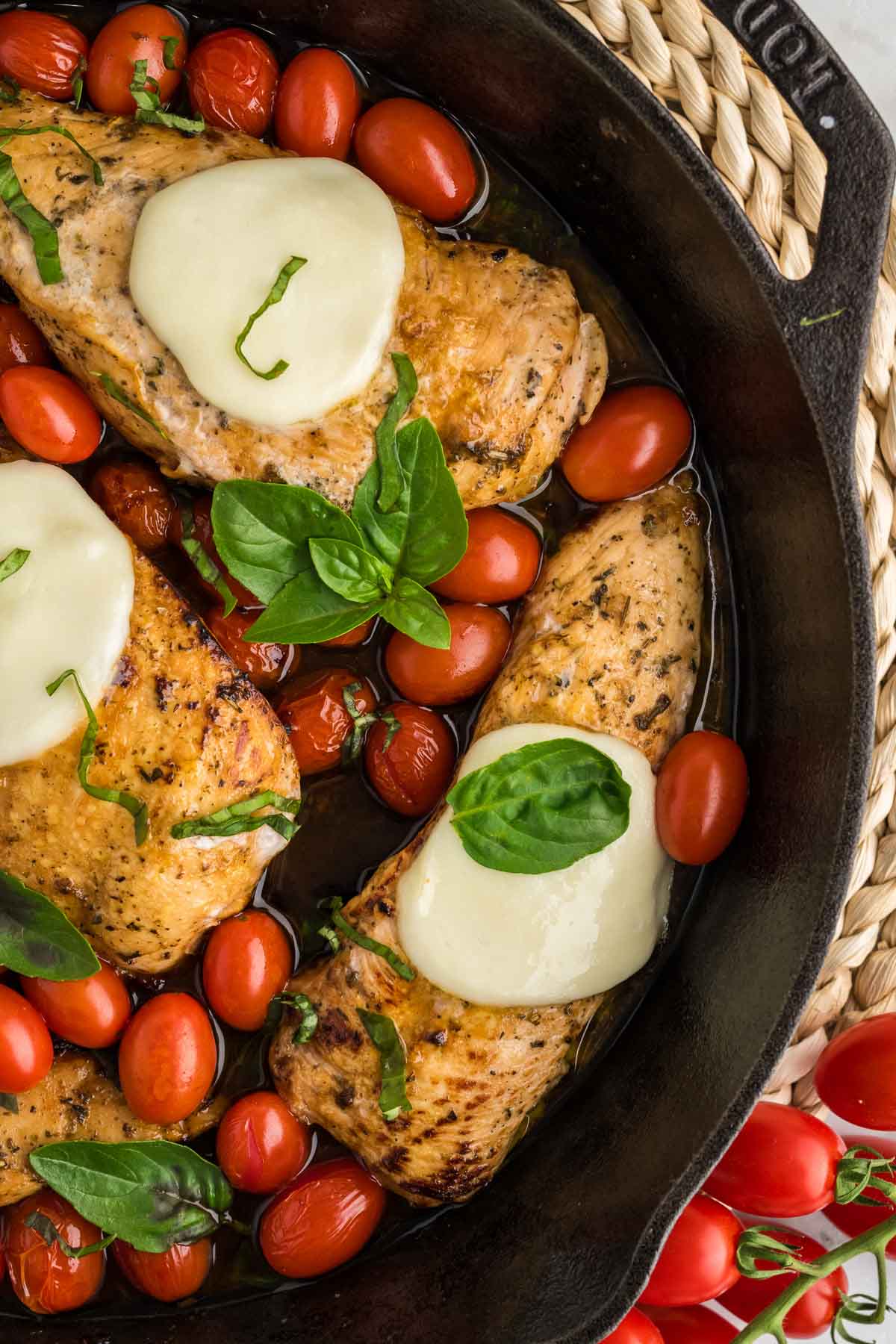 Three cooked chicken breasts topped with melted mozzarella cheese, cherry tomatoes, and fresh basil are arranged in a black skillet.