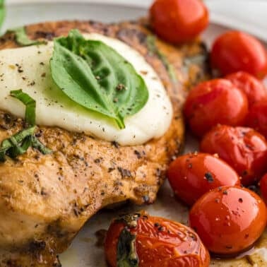 Grilled chicken breast topped with melted cheese and fresh basil, served with seasoned roasted cherry tomatoes and green beans on a plate.